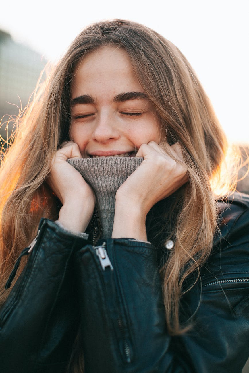 cheerful young woman with eyes closed covering mouth with turtleneck on street